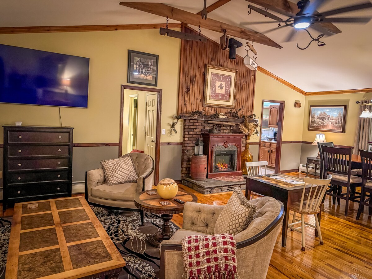 A welcoming living area features a large flat-screen television mounted above a rustic brick fireplace. Comfortable seating is provided by a pair of armchairs and a sofa. A wooden dining table sits nearby, surrounded by matching chairs, contributing to a relaxed atmosphere.