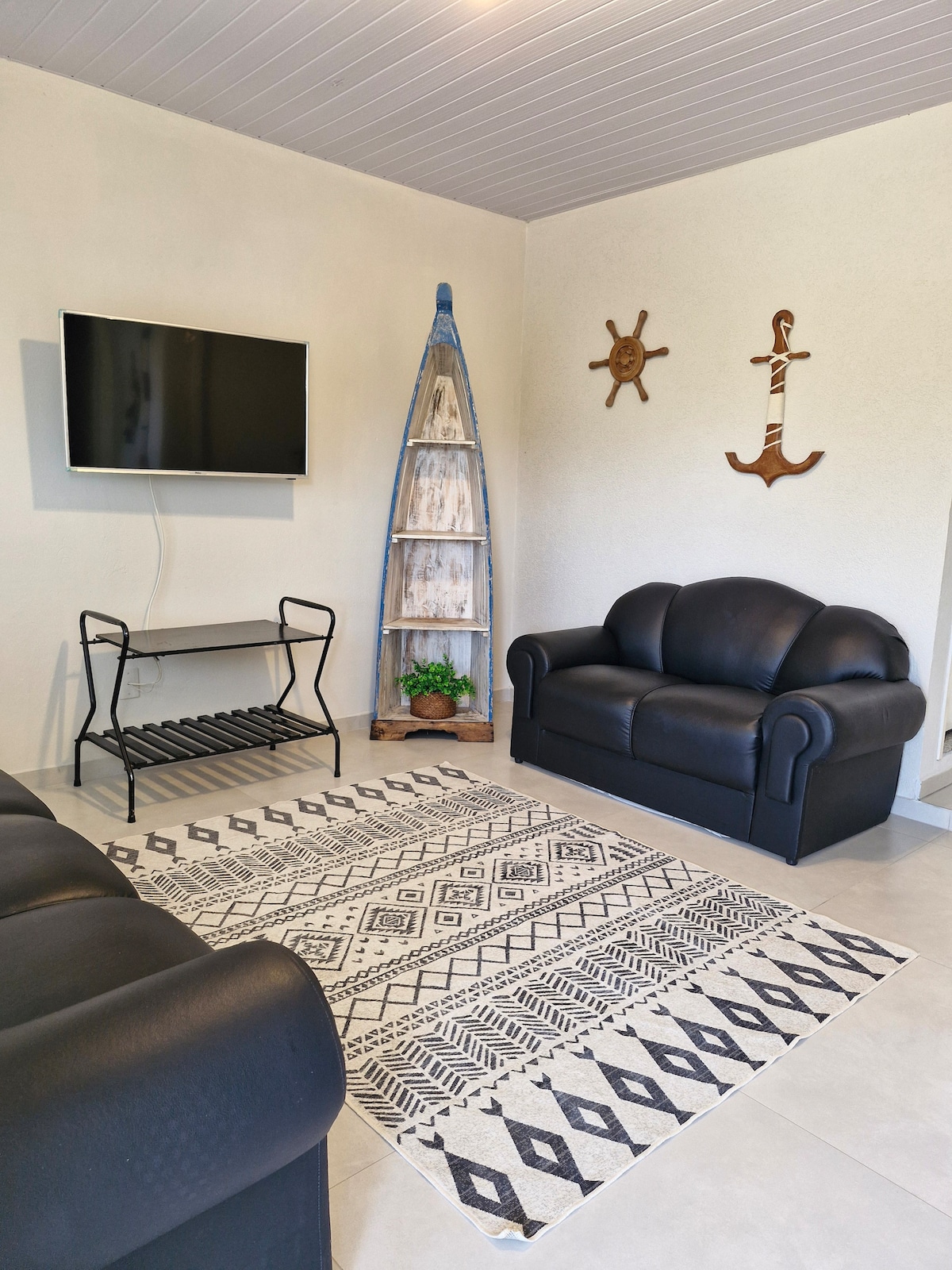 A cozy living area features two black sofas facing a mounted television. A decorative surfboard and nautical-themed wall art add character, while a patterned rug enhances the space. Natural light fills the room through a nearby window.