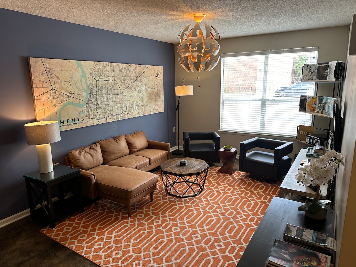 A cozy living area is showcased, featuring a brown sectional couch and two black armchairs. A round table sits at the center, complemented by an orange geometric area rug. A large map of Memphis adorns the wall, and natural light filters through the window.