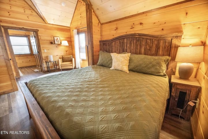 Recámara 1 | Nivel superior
Cama tamaño king con cabecera de madera oscura y ropa de cama verde. Mesita de noche con lámpara a la derecha, sala y ventanas visibles al fondo.