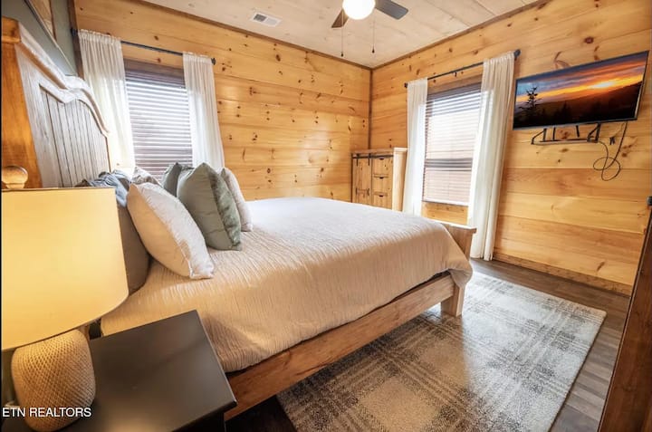 Recámara 2 | Planta principal
Cama tamaño king con plataforma, dos ventanas, cortinas transparentes, TV de pared y ventilador de techo. Puerta de granero visible al fondo. Paredes de pino nudoso en toda la propiedad.