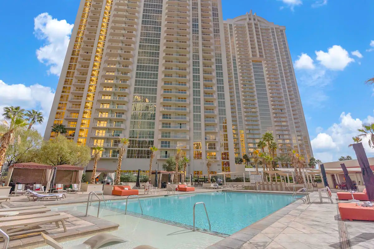 A serene pool area is surrounded by lounge chairs and palm trees, creating a relaxing atmosphere. Tall buildings reflect sunlight, while an inviting pool invites guests to unwind. Cabanas are positioned nearby for shade and comfort.