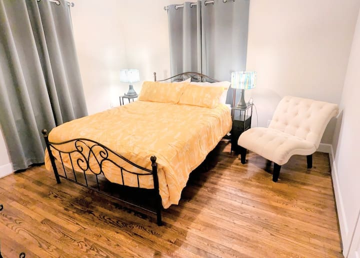 Dormitorio soleado con cama de hierro, pisos de madera y diván acolchado.