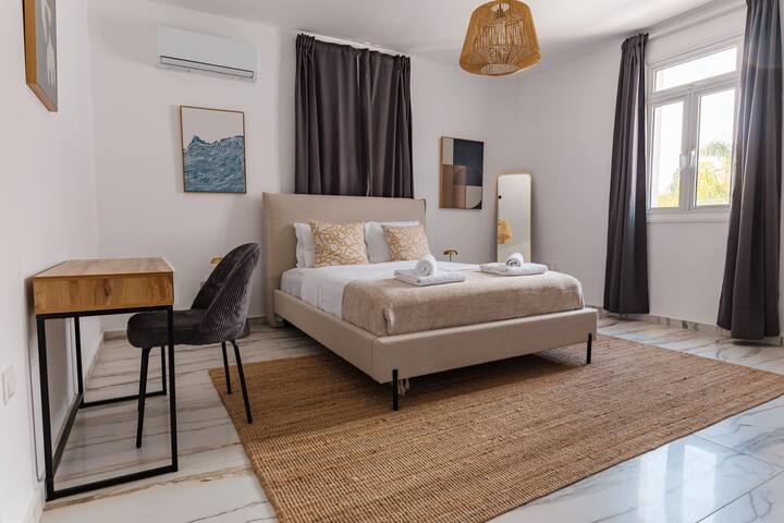 Acogedor dormitorio con ropa de cama en tonos neutros, mesita de noche de madera y alfombra de yute natural. Las amplias ventanas dejan entrar la luz natural y una lámpara colgante proporciona una iluminación suave.