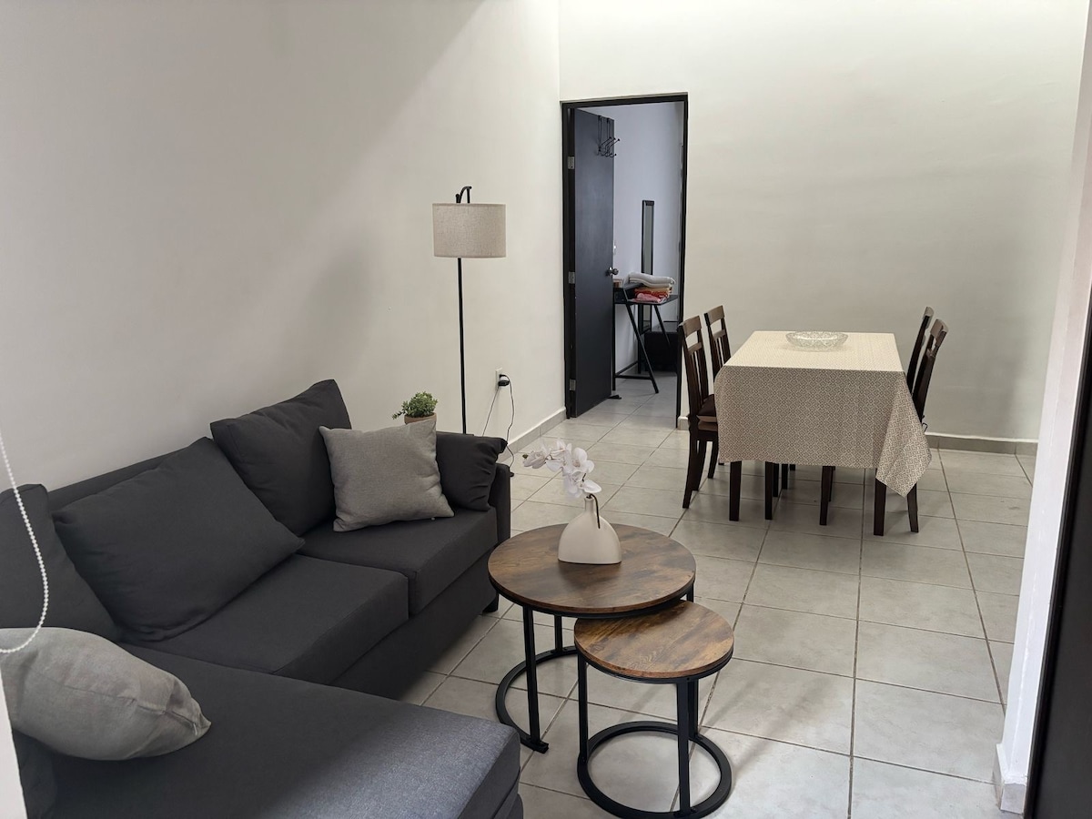 A spacious living area features a dark gray sectional sofa with decorative pillows. A small potted plant and a sculptural vase sit on two circular tables. A dining table set for six is positioned near a doorway leading to another room, complemented by warm lighting from a floor lamp.