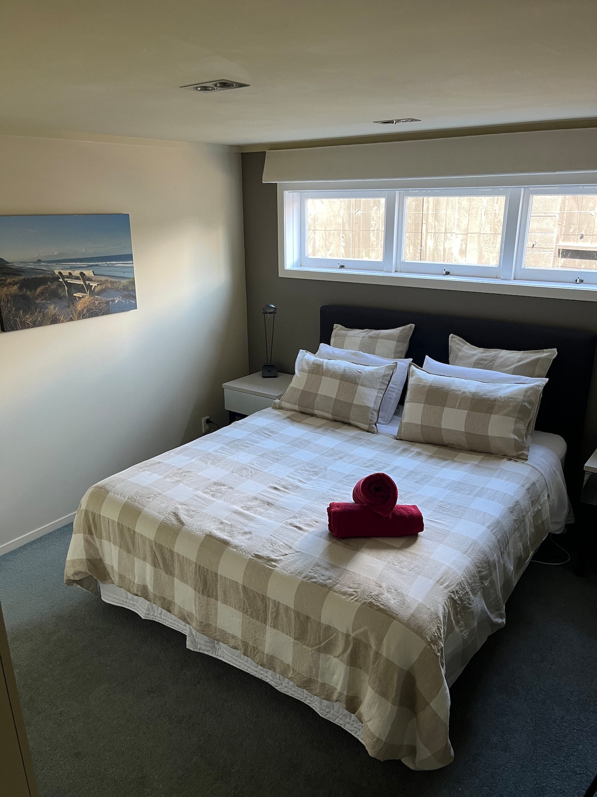 A cozy bedroom is presented with a spacious bed featuring a checkered comforter and decorative pillows. A neatly rolled towel is placed on the bed. Natural light filters through three narrow windows, providing a serene atmosphere.