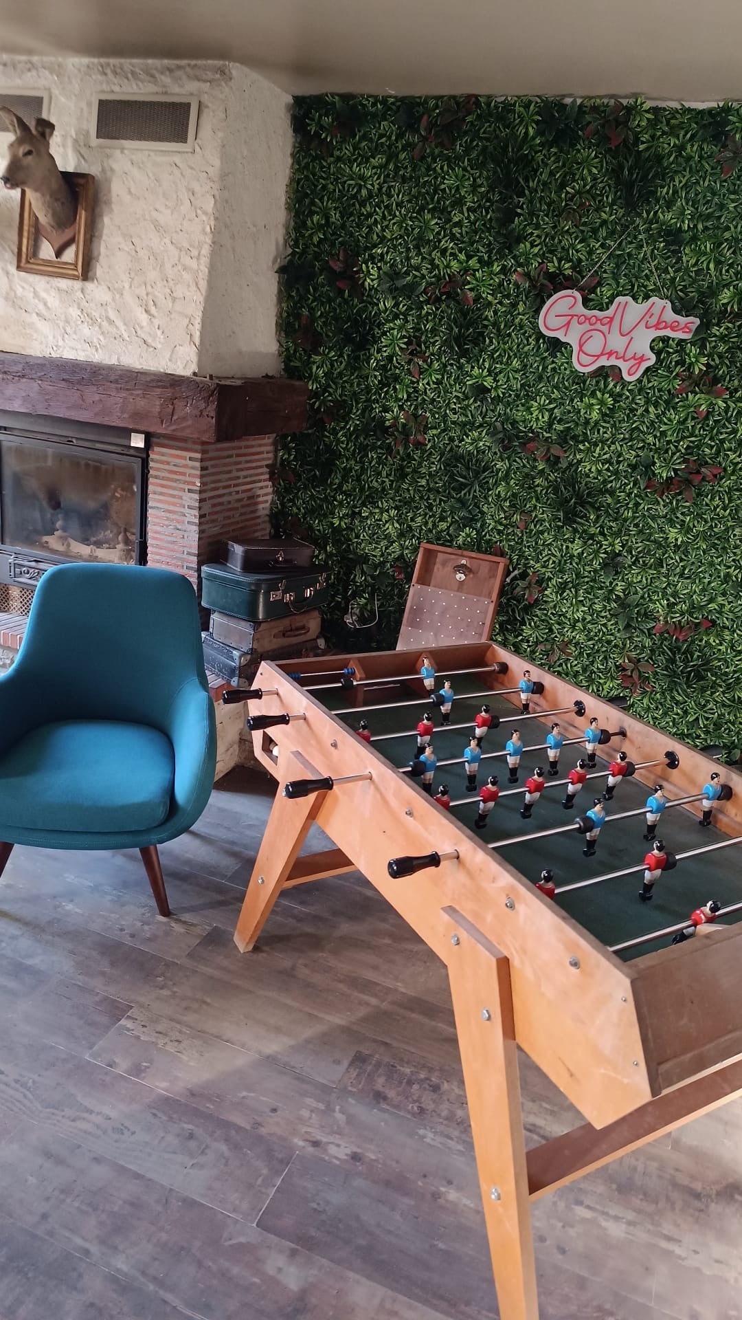 A wooden foosball table occupies a corner of the room, complemented by an upholstered chair in teal. A rustic fireplace, set with a stone mantel, is positioned against a backdrop of greenery. The wall features a sign that reads 'Good Vibes Only'.