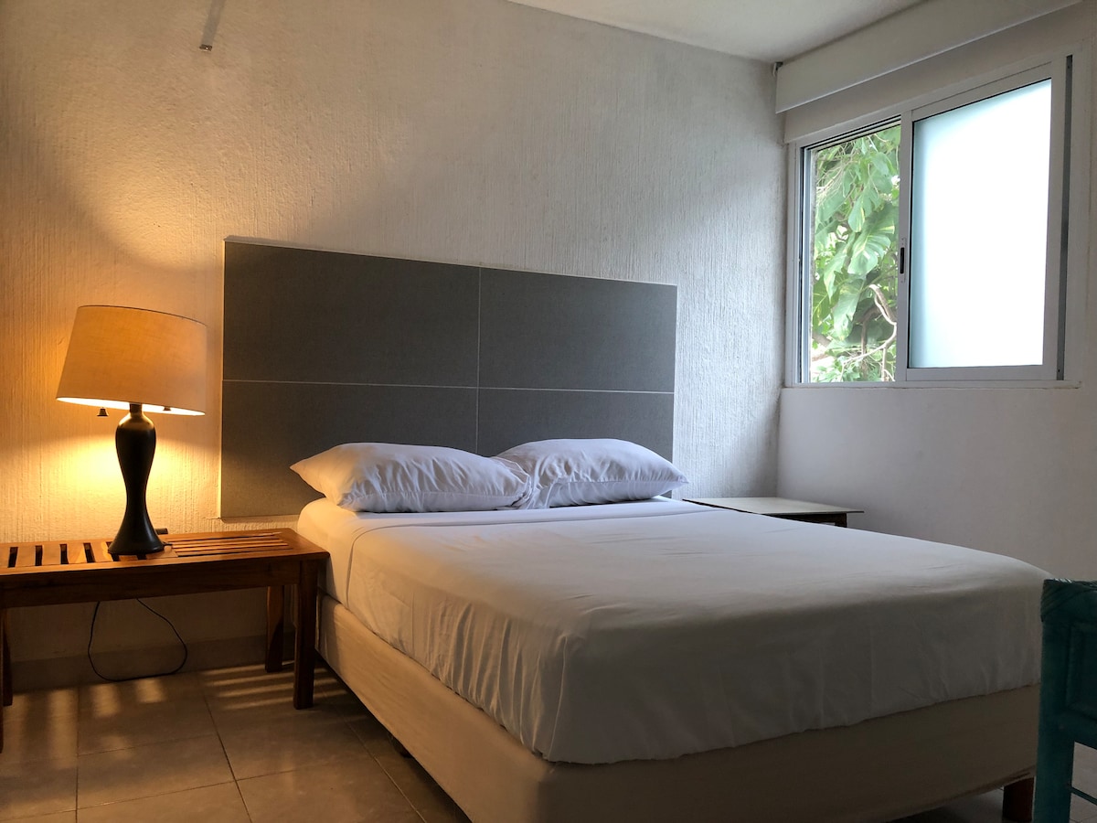 A well-lit sleeping area features a spacious bed dressed in crisp white linens. A bedside table supports a soft-glow lamp. A window allows natural light to filter in, complemented by a simple, neutral color palette on the walls and serene surroundings visible outside.