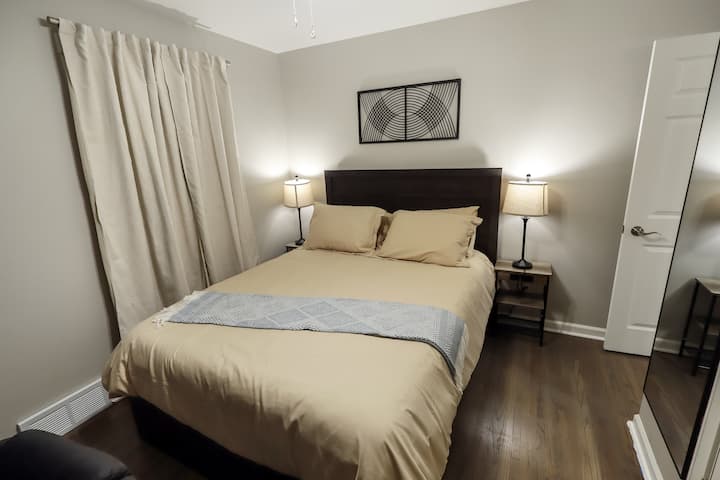 Cama cómoda tamaño queen con sábanas de colores neutros, dos lámparas de noche y un gran espejo de pared. Las paredes de color gris suave y la luz natural crean un refugio tranquilo y acogedor.  Ubicado en el nivel principal.
