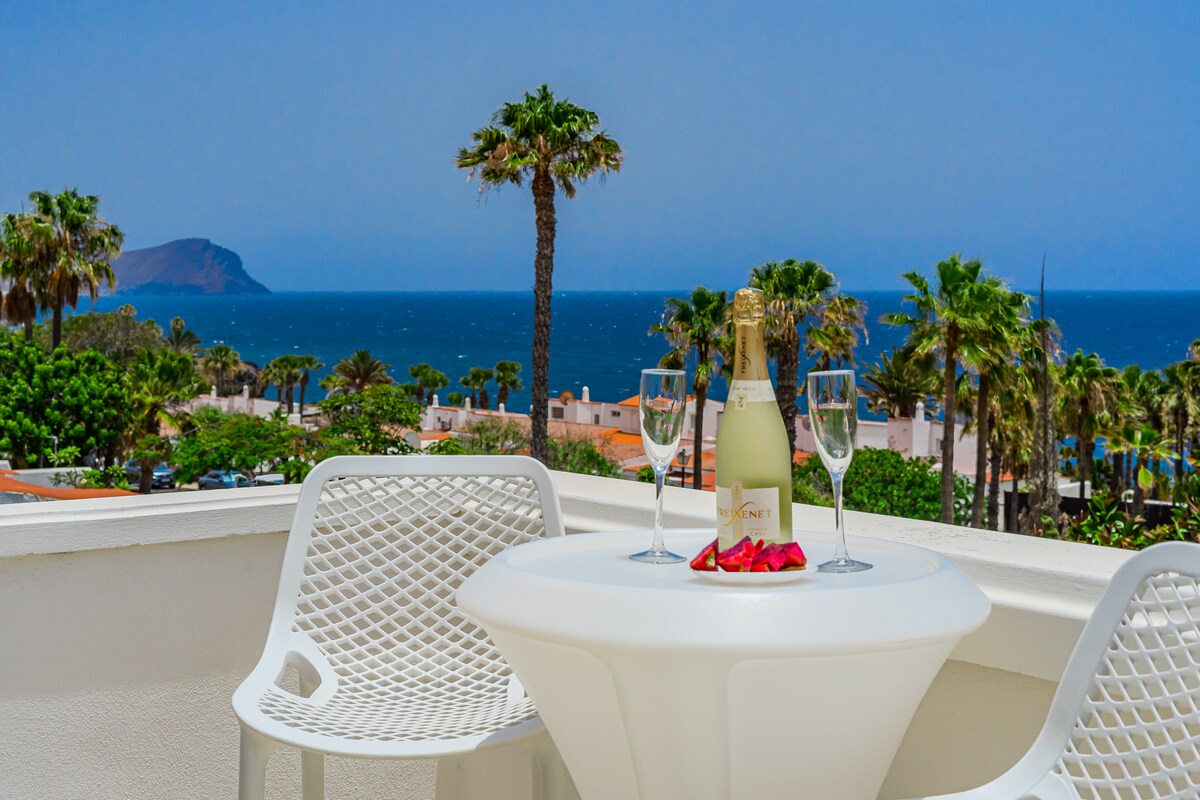 A white bistro table is set on a terrace, accompanied by two glasses and a bottle of champagne. Lush palm trees frame the background, with a panoramic view of the Atlantic Ocean and distant island visible under a clear blue sky.