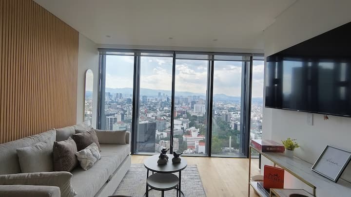 Sala elegante con ventanales de piso a techo y espectacular vista panorámica de la Ciudad de México, ideal para una estancia premium en Reforma.