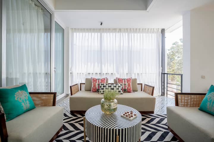 A sunlit sitting area with an open, airy feel and a modern aesthetic. Light-toned sofas and armchairs with woven wooden frames are arranged around a round coffee table, placed on a bold black-and-white patterned rug.