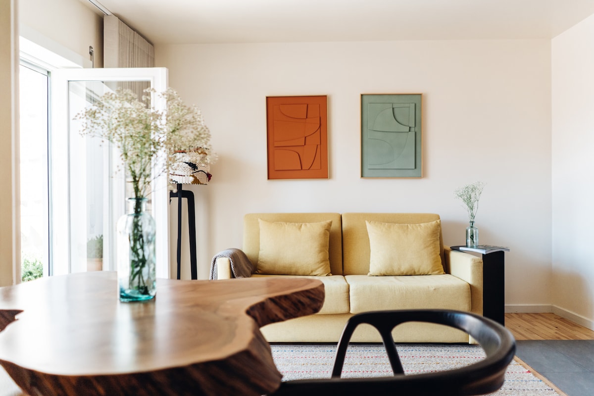 A bright living area features a yellow sofa and two abstract wall art pieces in warm and cool tones. A wooden table with natural edges adds texture, while vases with flowers enhance the space's charm. Light pours in through the open doors, connecting to the outdoors.