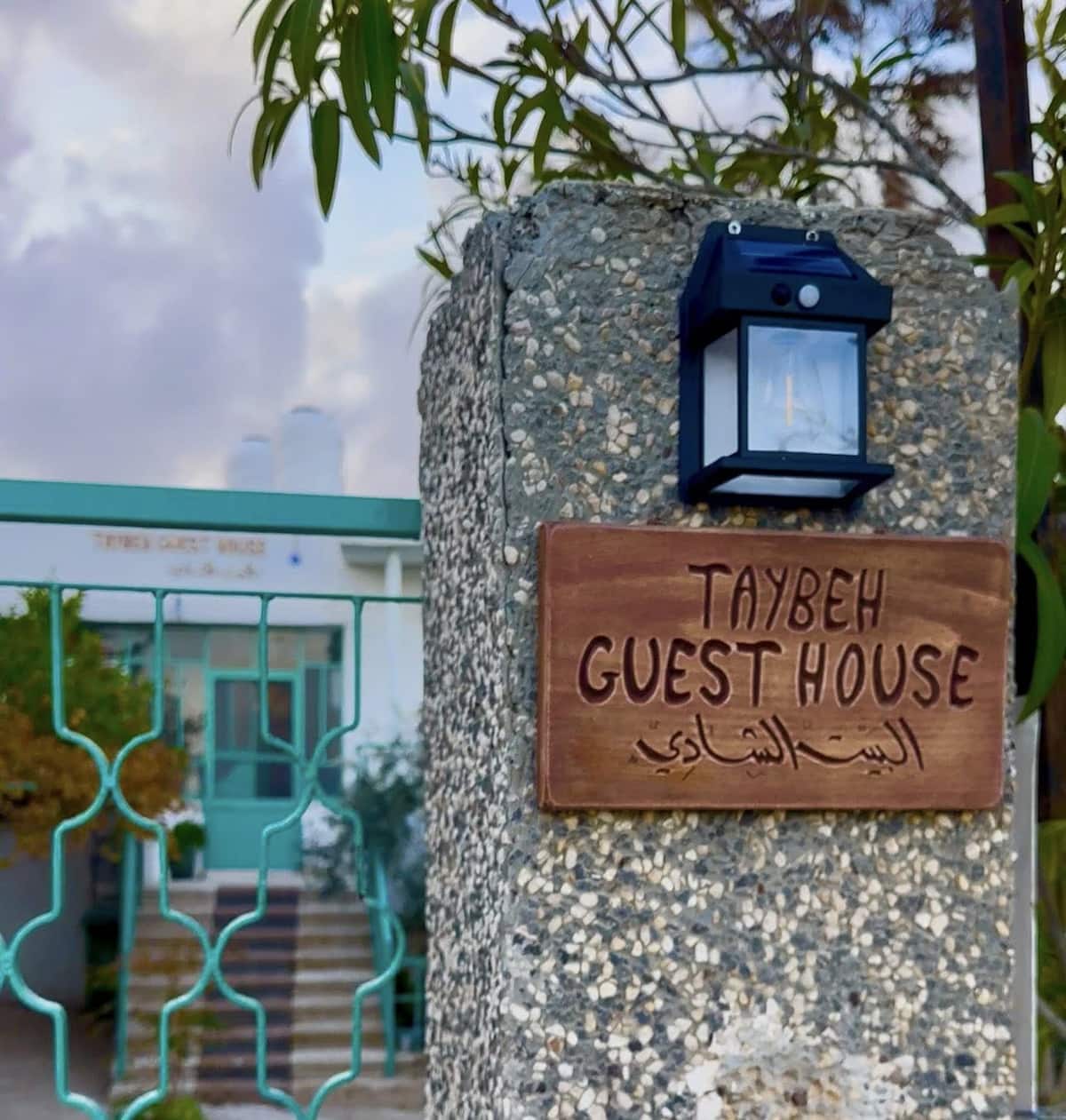 Carved wooden Taybeh Guest House sign on a stone gate pillar beneath a lantern