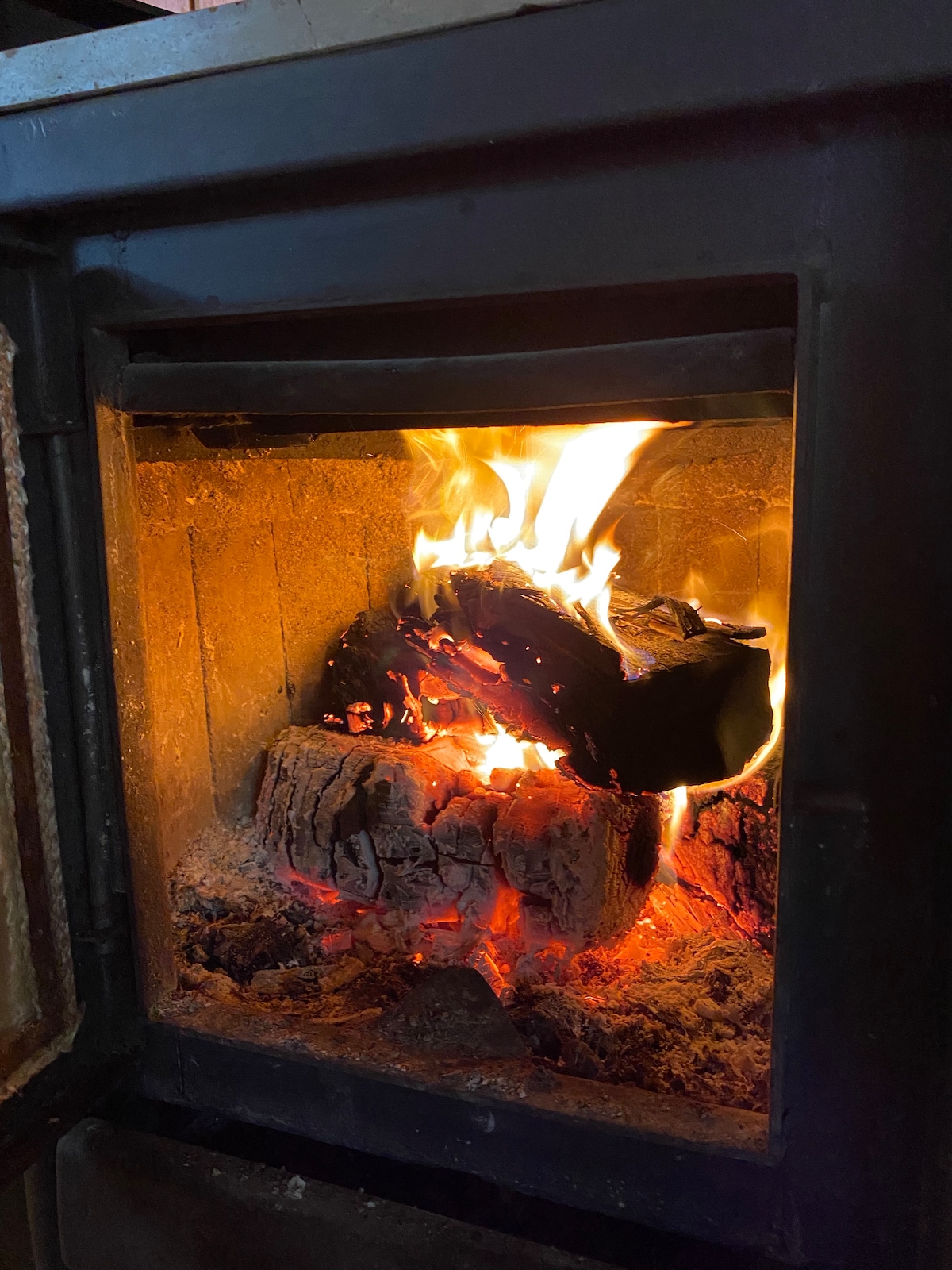 A wood-burning stove emits a warm glow, with flames flickering above logs and embers. The textured surface of the stove is visible, highlighting the rustic charm. The light from the fire adds warmth to the area around it.