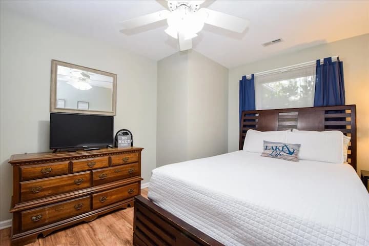 Habitación para huéspedes con cama queen bañada por el sol, cama de madera color miel, cortinas azul marino y un sofá cama adicional. Cama queen, decoración con temática lacustre, televisor de pantalla plana, paredes grises, almohada con motivos costeros, piso de madera flotante, escapada a Smith Mountain Lake.