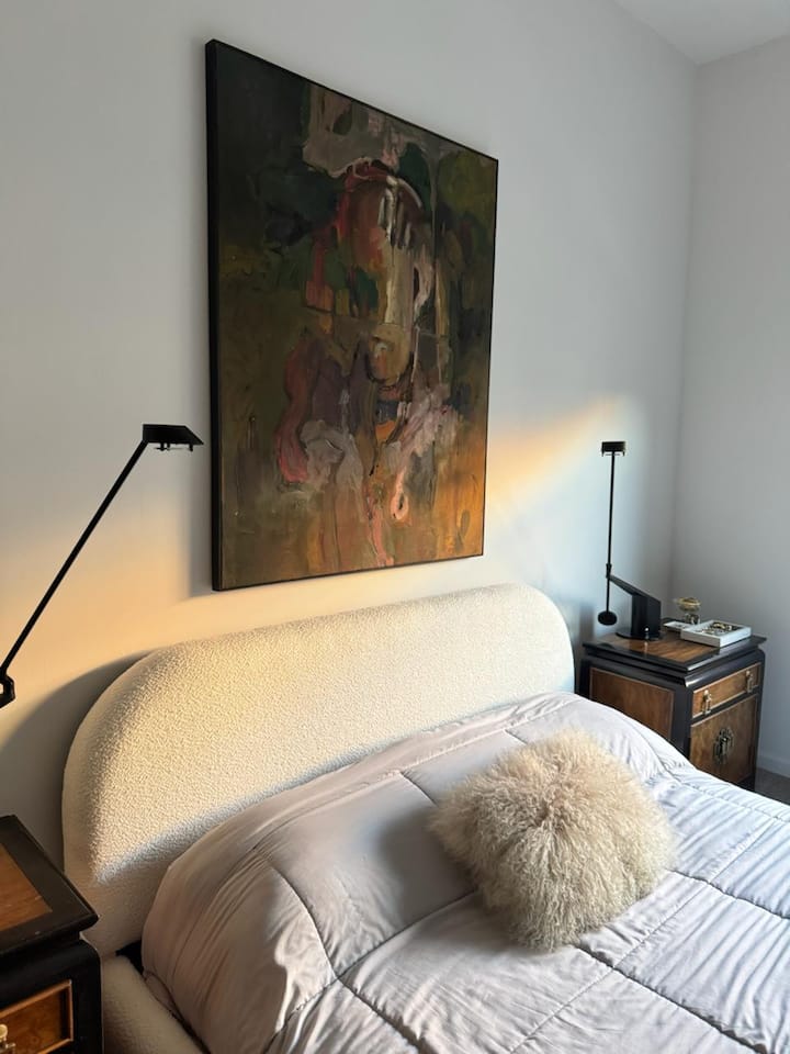 Cozy bedroom corner with a plush upholstered headboard, soft neutral bedding, and warm ambient lighting. A statement art piece adds character, creating a calm and inviting space to relax and unwind.
