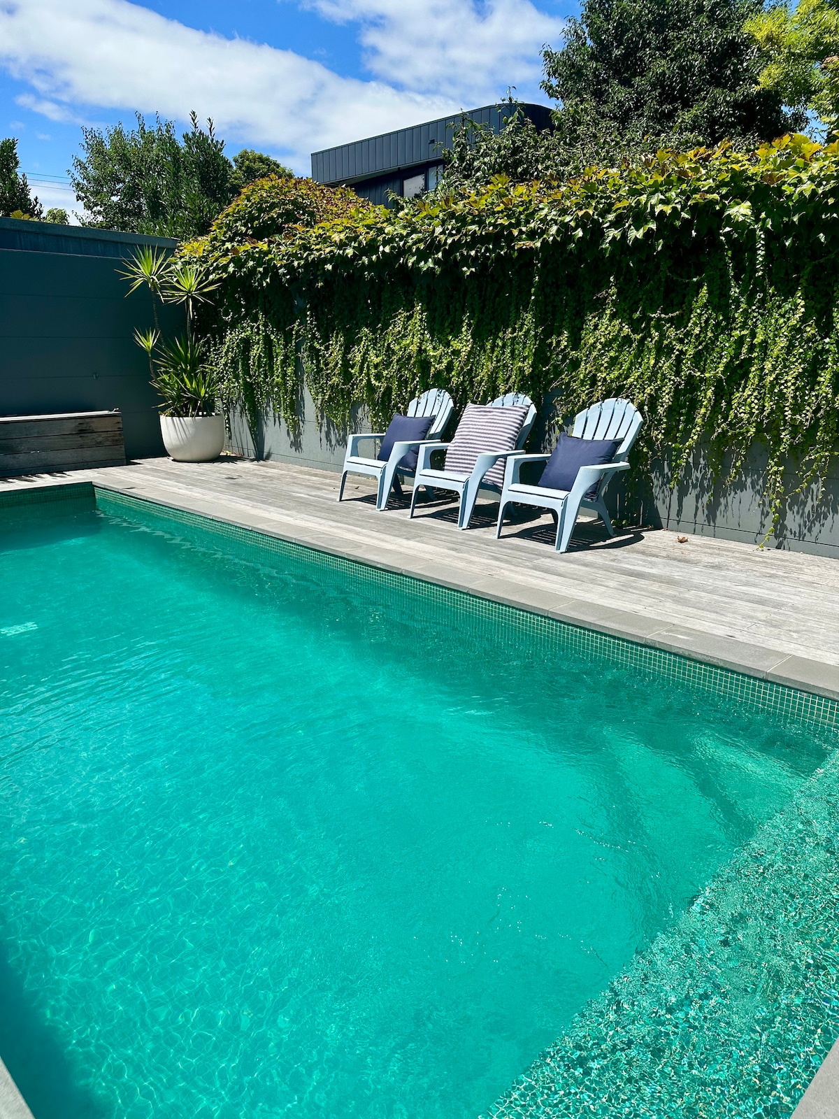 A serene pool area is featured, showcasing clear turquoise water surrounded by wooden decking. Several blue and white striped chairs are positioned nearby, complemented by lush greenery along the wall, creating a tranquil outdoor ambiance.