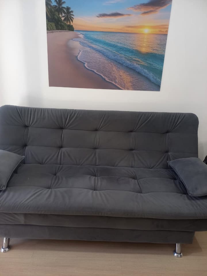 Un salón acogedor y moderno con un cómodo sofá, ideal para relajarse después de un día de turismo. Un ambiente limpio y climatizado, perfecto para momentos de descanso, con una vista decorativa inspirada en una puesta de sol en la playa. ✨