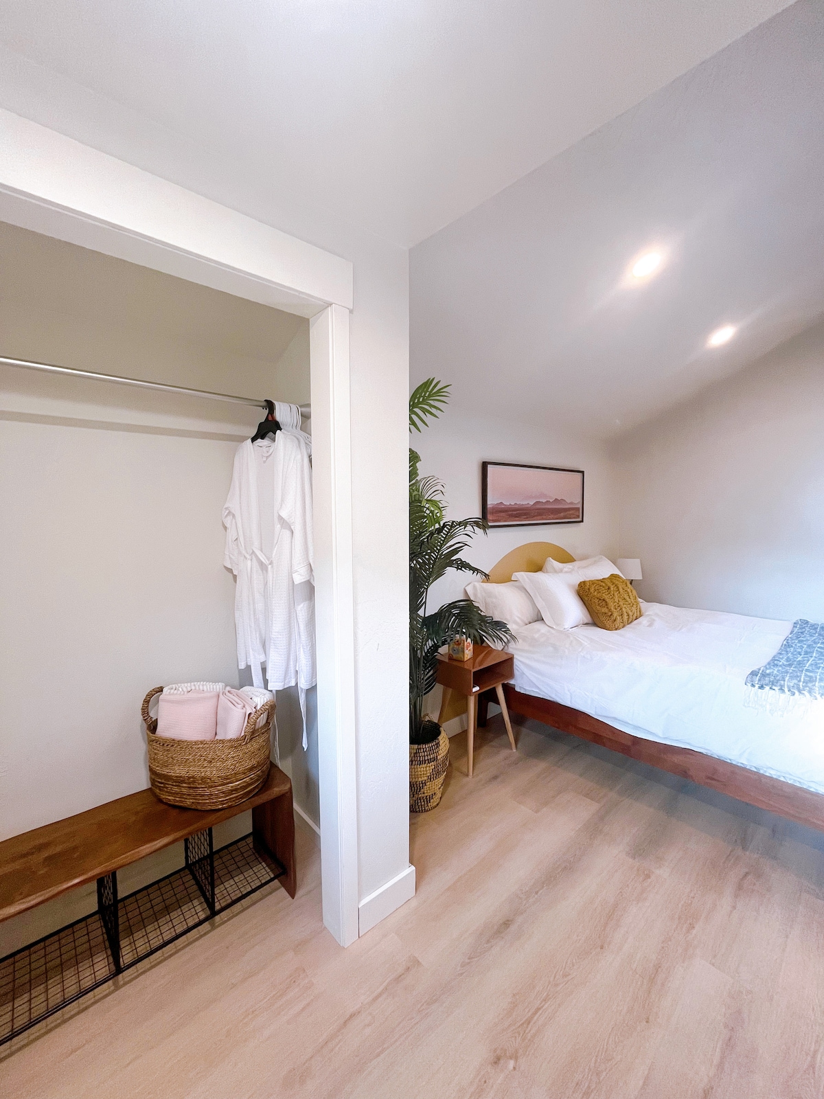A cozy bedroom is showcased, featuring a spacious bed with neutral linens and decorative pillows. A small bench with a basket holding folded towels is positioned near the entryway. A hanging rod with garments can be seen in the closet area, along with a plant adding a touch of greenery.