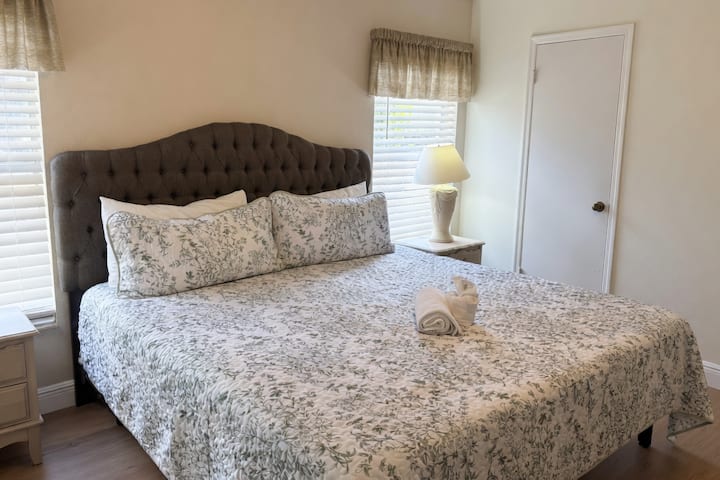 Recámara principal relajante con una cama king grande, cabecera acolchada y elegante ropa de cama con estampado floral. Incluye una mesita de noche y una lámpara para mayor comodidad.