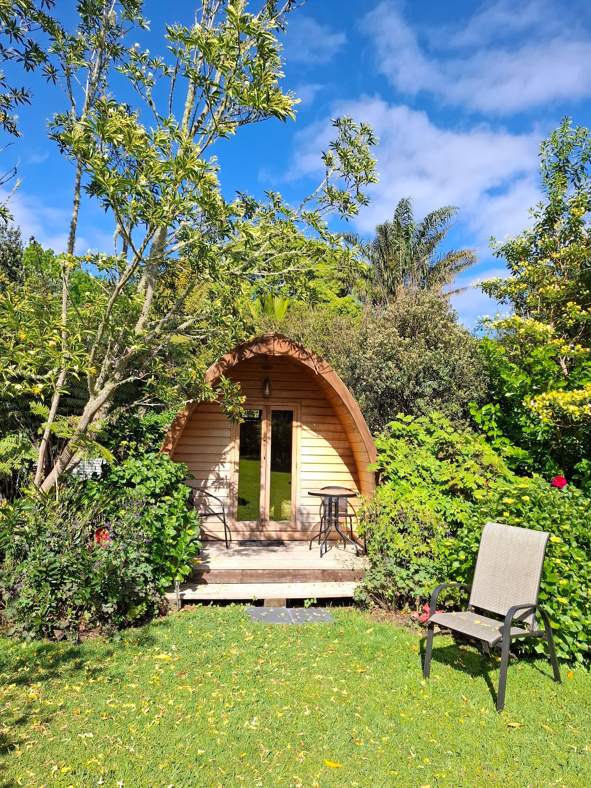 An EcoPod is nestled amongst lush greenery, featuring a wooden exterior with a curved roof. A small porch with two chairs invites relaxation, while tall trees and vibrant plants enhance the natural surroundings under a bright blue sky.