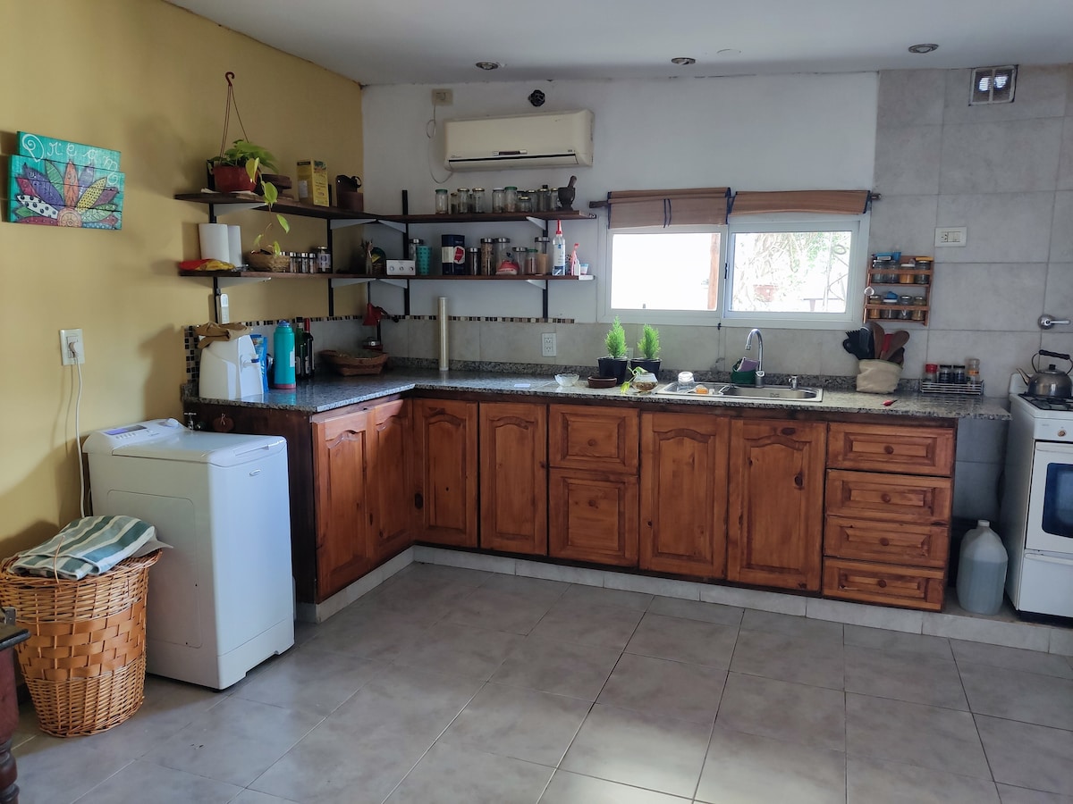 A spacious kitchen is featured with wooden cabinetry and a granite countertop. Open shelves display various jars and utensils. Natural light enters through two windows, while a washing machine is positioned next to the sink. A cozy, inviting atmosphere is evident in the layout.