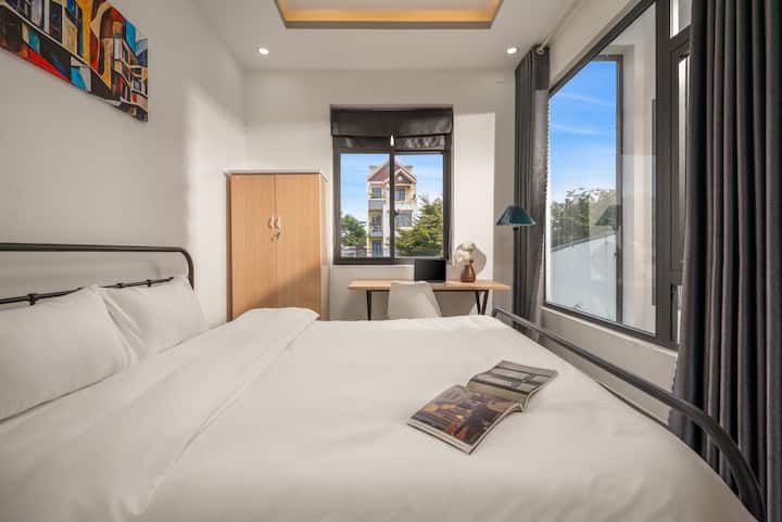 Dormitorio minimalista con una cama de hierro negro y ropa de cama de un blanco inmaculado. El área de trabajo está ubicada junto a una gran ventana, lo que proporciona luz natural y una vista abierta a la calle. El mobiliario incluye gabinetes de madera de color claro y lámparas de pie modernas, lo que crea un espacio relajante y cómodo para descansar.