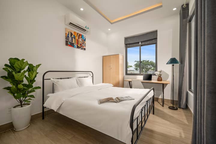 Un dormitorio moderno con una distintiva cama de marco de hierro negro, que crea un punto focal contra una pared blanca. El espacio está totalmente equipado con un escritorio junto a la ventana, un armario de madera y plantas decorativas, lo que crea un ambiente juvenil, espacioso y lleno de energía.