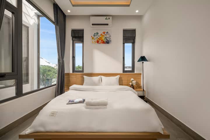Un dormitorio de estilo minimalista con una cama doble de color blanco nítido y una elegante cabecera de madera. El gran sistema de puertas de vidrio ofrece vistas abiertas, combinadas con pinturas artísticas en las paredes y lámparas de pie modernas para crear un espacio de descanso acogedor y lleno de luz.