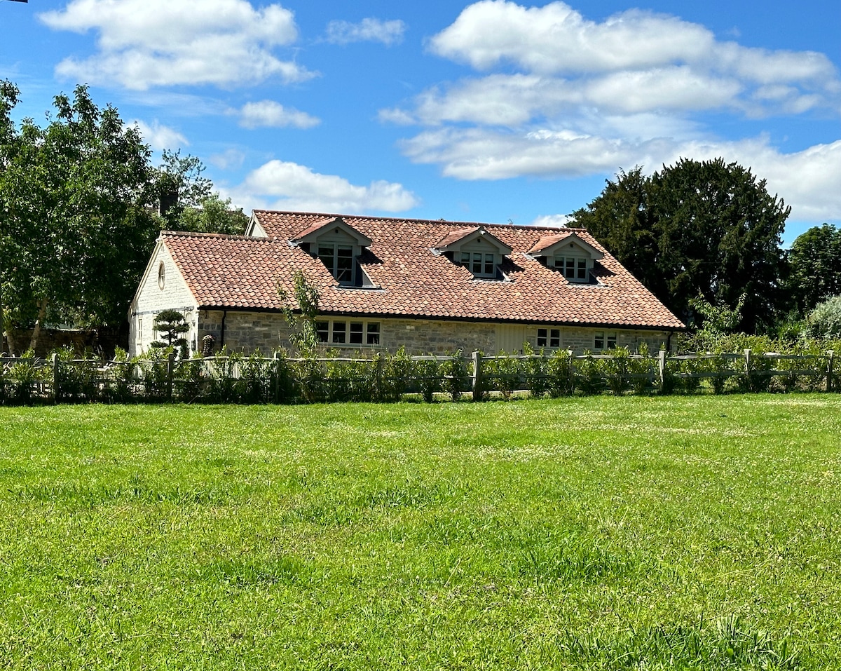 A spacious barn conversion is set within lush green paddocks, featuring a charming stone exterior and a red-tiled roof. Three dormer windows provide natural light, and the surrounding landscape showcases vibrant grass under a partly cloudy sky.