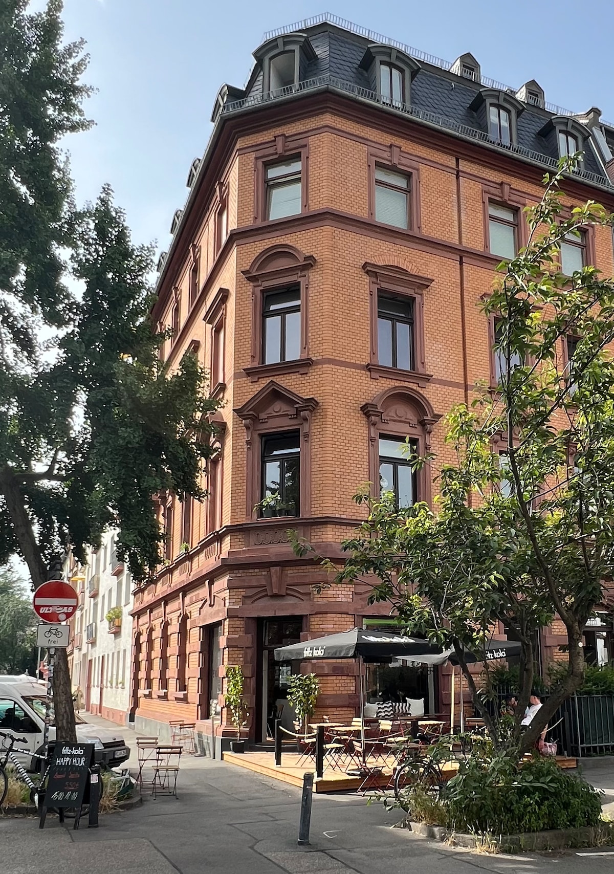 A historical building showcases a classic red brick facade with intricate architectural details. Paned windows reflect natural light, while outdoor seating areas are set beneath canopies. Lush greenery frames the entrance, contributing to the inviting ambiance of the vibrant street scene.