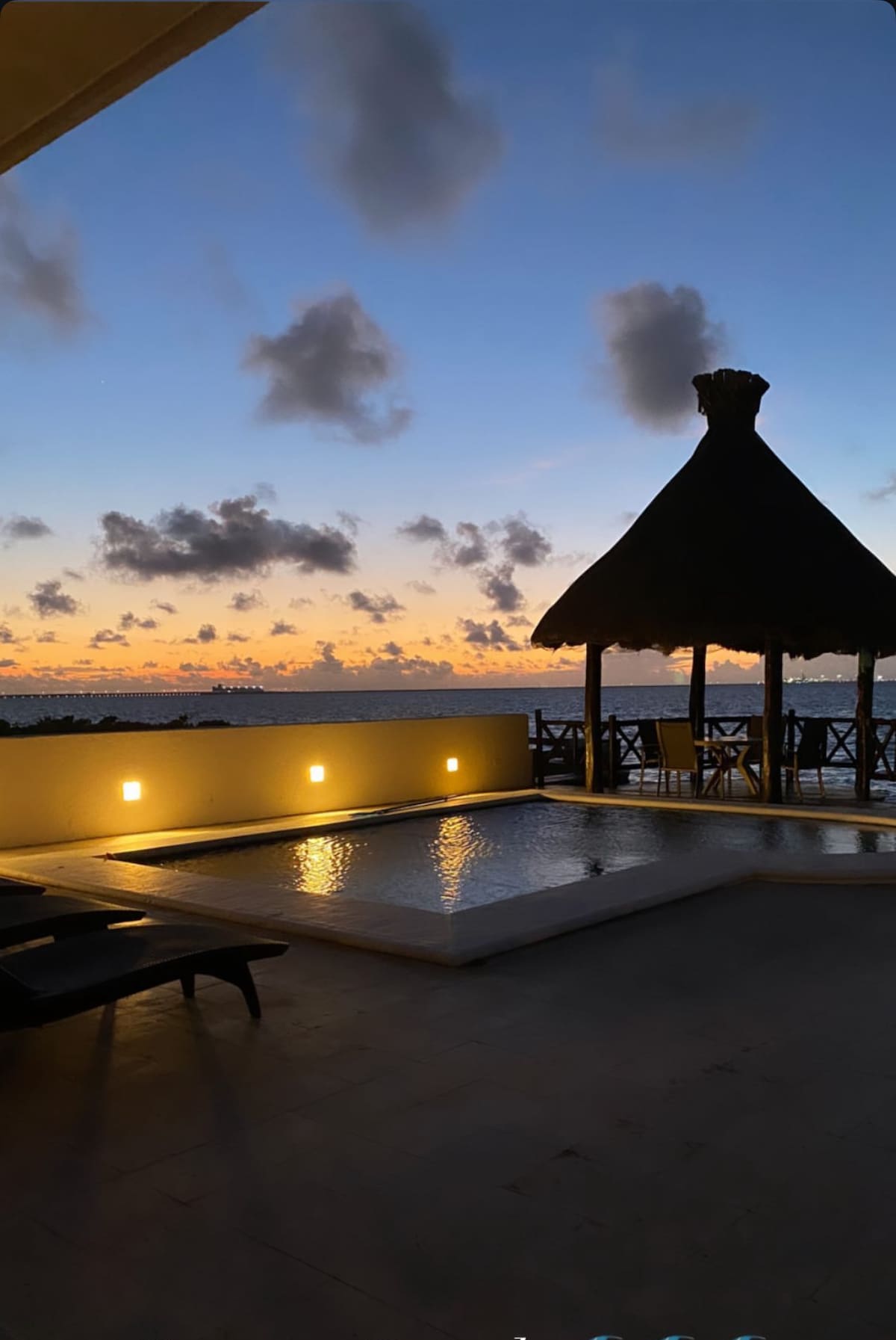A serene outdoor space is highlighted by a swimming pool reflecting the evening sky's colors. A shaded gazebo with tall palm leaves provides seating. The silhouette of distant clouds enhances the tranquil beachfront atmosphere as the sun sets over the ocean.