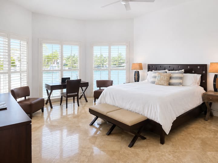 Bright and spacious master bedroom with a king bed, sitting area, desk, and beautiful water views through large plantation-shuttered windows.