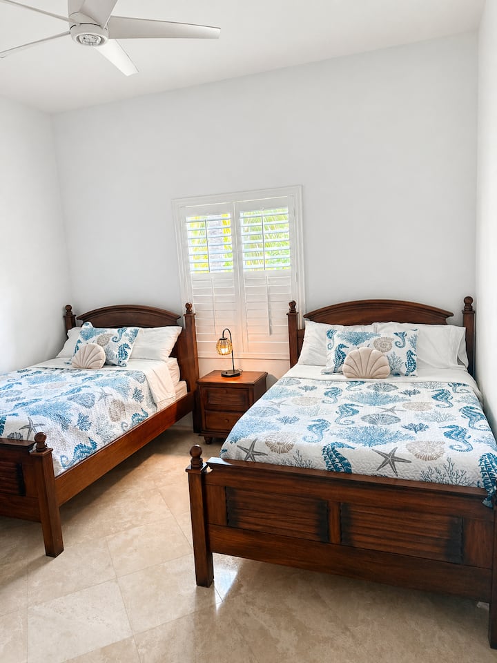 Coastal bedroom with two double beds, beach-inspired bedding, ceiling fan, and bright natural light — perfect for kids, friends, or additional family guests.
