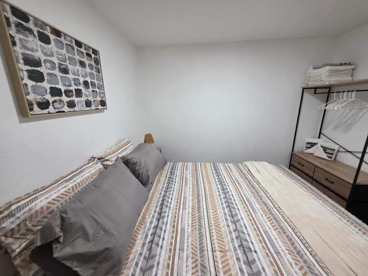 ”Rum med en dubbelsäng, sängkläder i neutrala nyanser och enkel inredning.” Inkluderar ett nattduksbord med en lampa och ett öppet skåp med hyllor och hängutrymme. Välstädat och väl upplyst utrymme.”