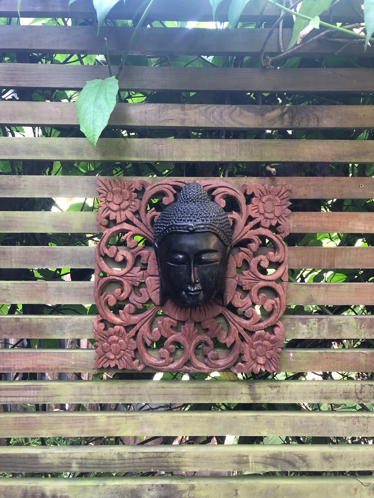 A decorative wall feature is displayed, showcasing intricate floral carvings in earthy tones. A minimalist black Buddha head is mounted at the center, surrounded by the ornate design, which adds a serene touch to the natural setting.
