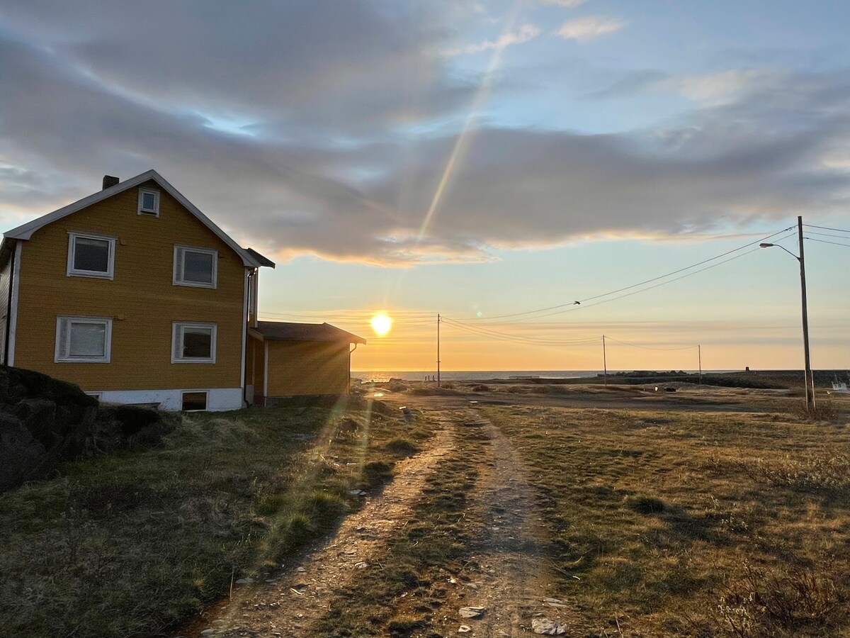 A yellow house is positioned beside a gravel path, with the sun setting on the horizon. Soft clouds diffuse the light, creating a warm glow over the grassland. Power lines run alongside the path, leading toward the distant coastline.