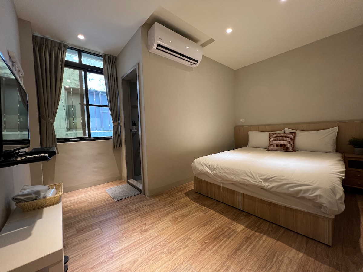 A clean and bright room is featured, highlighting a spacious bed dressed in soft linens. A window allows natural light to flood the area, complemented by light-colored walls and wooden floors. An air conditioning unit is mounted on the wall for added comfort.
