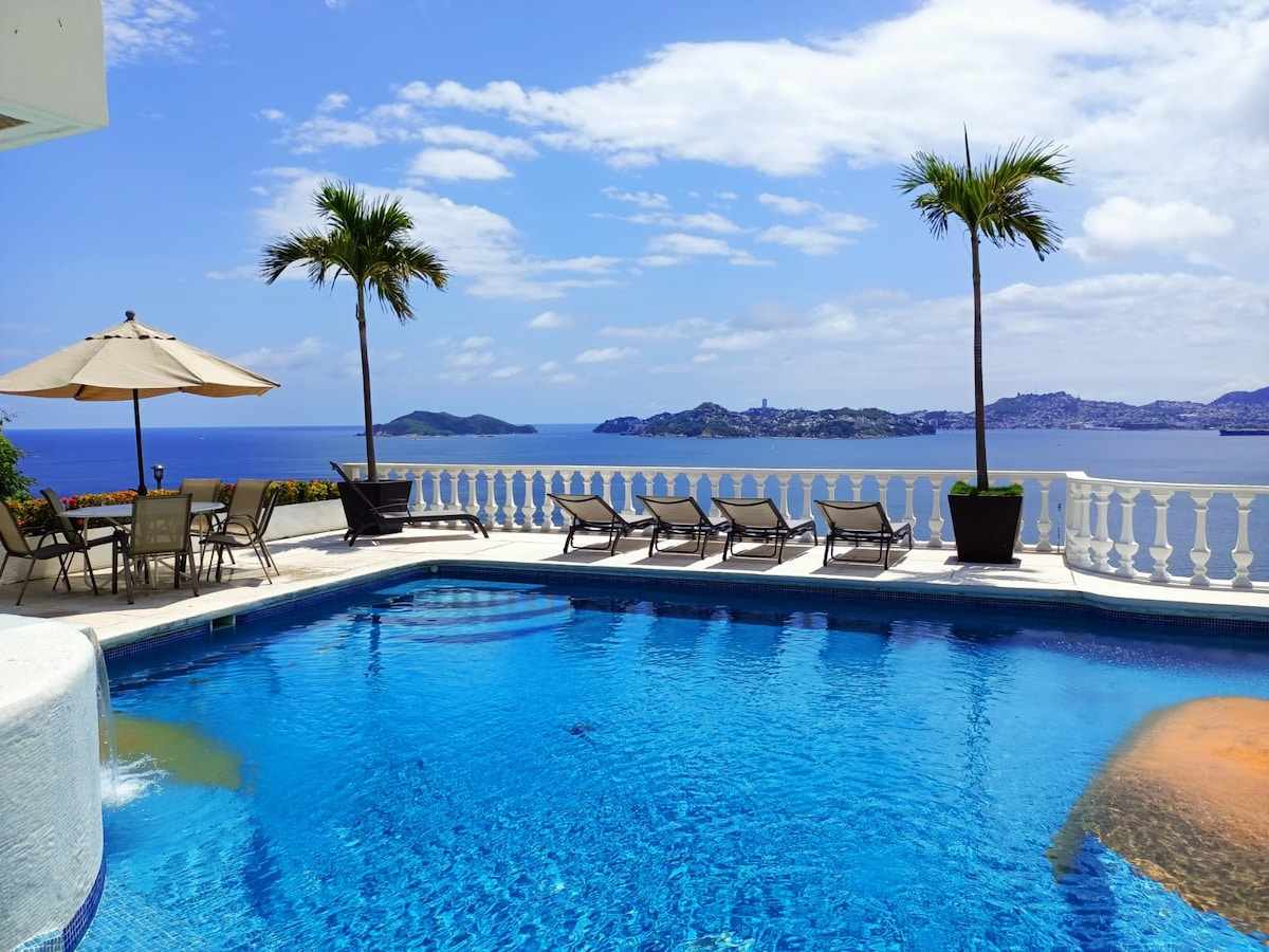 A clear blue pool reflects the sky and surrounding landscape. Several lounge chairs are positioned near the water's edge, providing space for relaxation. A shaded area with an umbrella and a table is visible in the background, accompanied by palm trees framing the scenic view of the ocean and hills beyond.