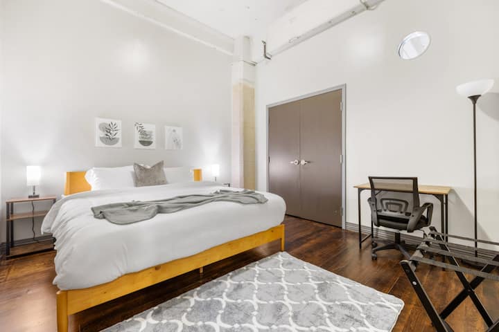 Moderna suite con dormitorio que cuenta con una cama de plataforma de color amarillo mostaza, suelos de madera, un sillón decorativo, TV montada en la pared y entrada al baño privado.