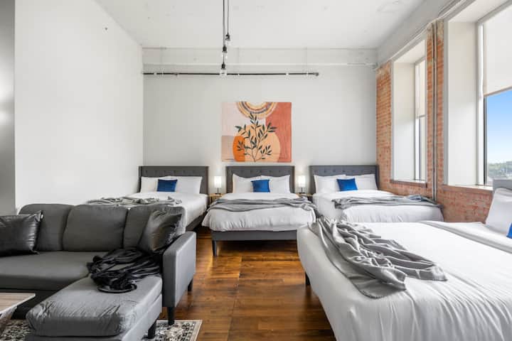 Estudio tipo loft de concepto abierto con cuatro camas tamaño queen, techos altos, paredes decorativas de ladrillo a la vista, suelo de madera noble y una zona de estar con sofá modular.