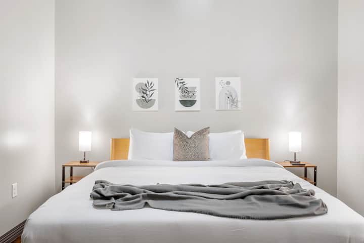 Dormitorio sereno con una cama tamaño king con sábanas blancas impolutas, cabecero de madera clara, mesitas de noche con lámparas a juego y arte mural minimalista.