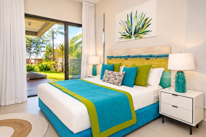 Lit double avec tête en bois naturel, coussins turquoise et vert, lampes de chevet bleues et tapis jute. Baie vitrée ouverte sur le jardin et la terrasse. Chambre lumineuse et colorée.