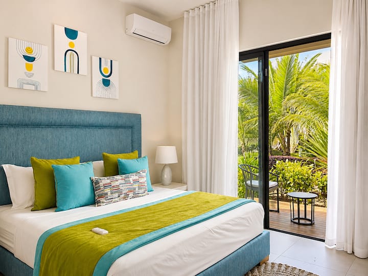 Chambre avec lit queen size, tête de lit bleue, coussins turquoise et vert anis. Climatisation, baie vitrée coulissante avec vue directe sur les palmiers et la terrasse extérieure.
