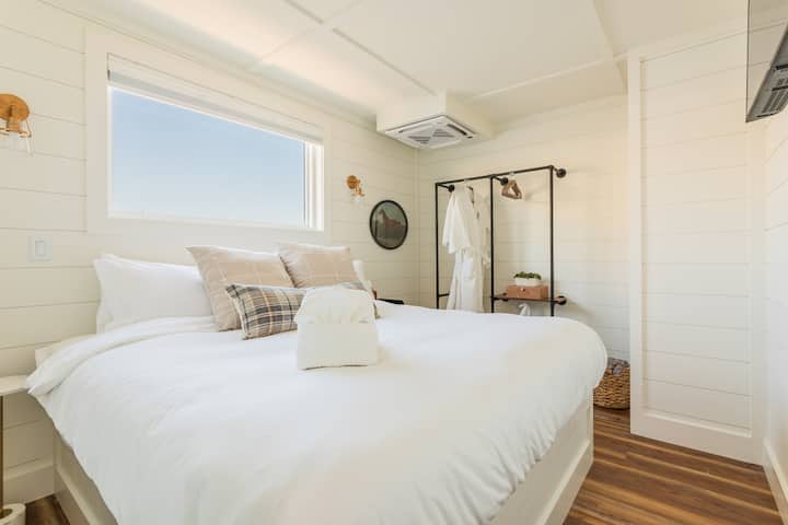La segunda suite privada King Master, decorada individualmente con cálidas sábanas a cuadros, paredes de tablas superpuestas, apliques de latón, una bata FLOHOM en el perchero del armario abierto y su propio baño privado justo al otro lado de la puerta.