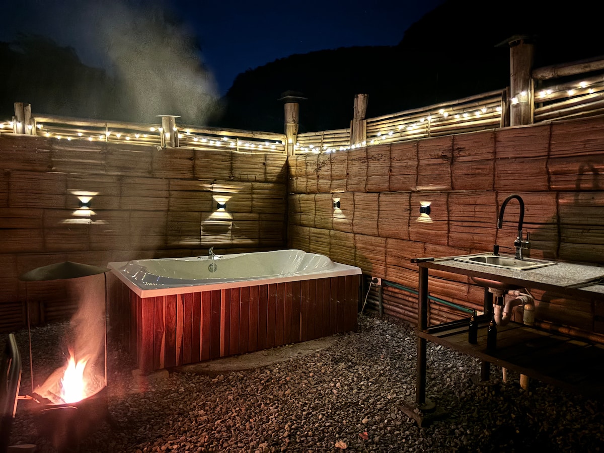An outdoor hot tub is surrounded by bamboo walls, softly illuminated by string lights overhead. A small fire pit flickers nearby, providing warmth. A functional sink area is visible in the background, contributing to the relaxing ambiance.