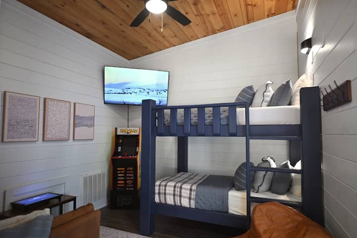 The Hideout Loft features built-in twin bunks with individual reading lights, a cozy lounge area, Smart TV, Infinity Game Table, and space to relax after a day of adventure.