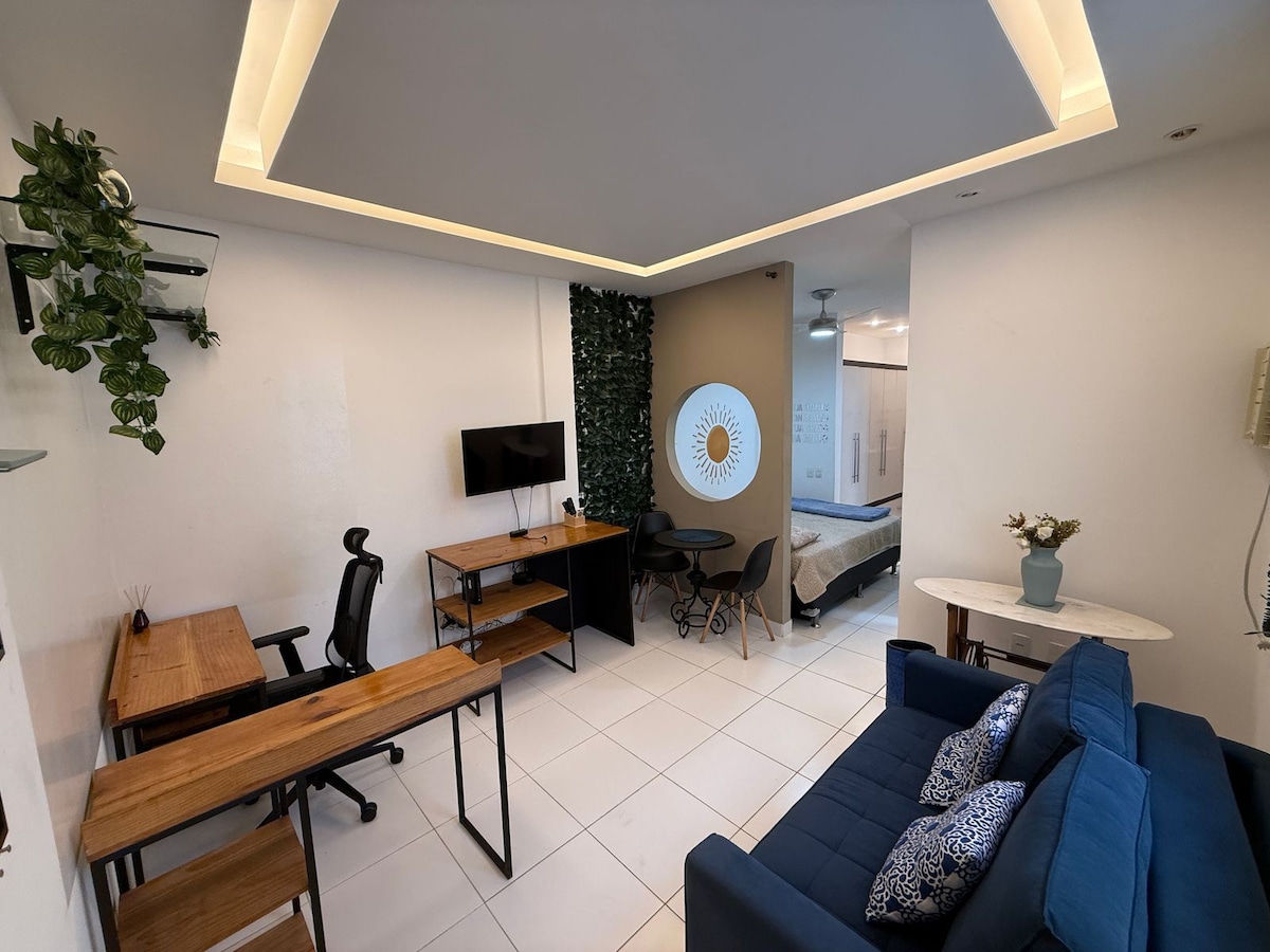 A bright living area is depicted featuring a blue sofa and a wooden desk with a comfortable chair. A wall-mounted television and decorative wall art add interest. The space showcases modern furnishings against a backdrop of greenery and light-colored walls, enhancing the inviting ambiance.