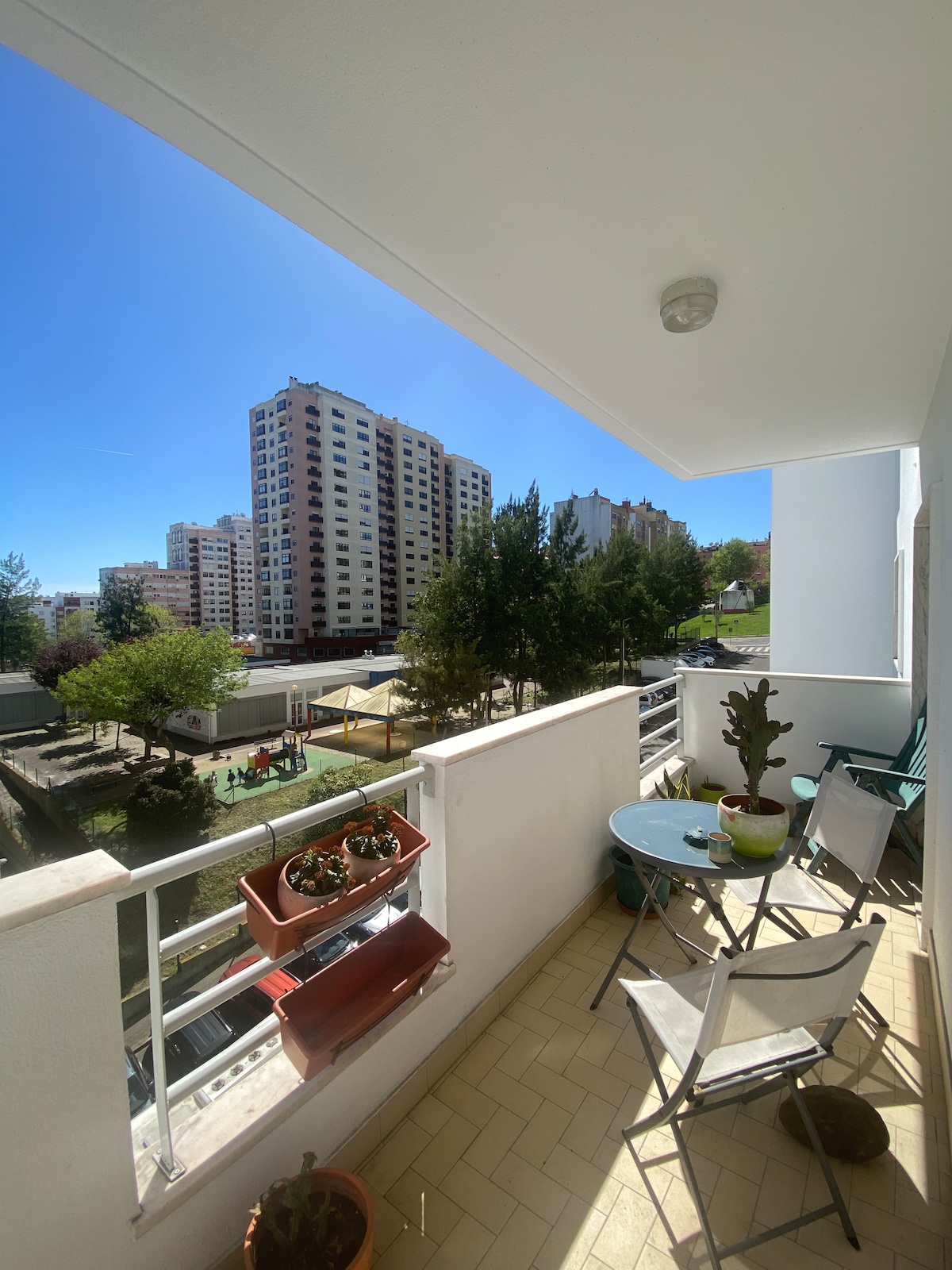 A private balcony is shown, furnished with a small round table and two chairs. Potted plants are placed on the table and a wall-mounted planter. A view of neighboring buildings and greenery is visible in the background, under a clear blue sky.